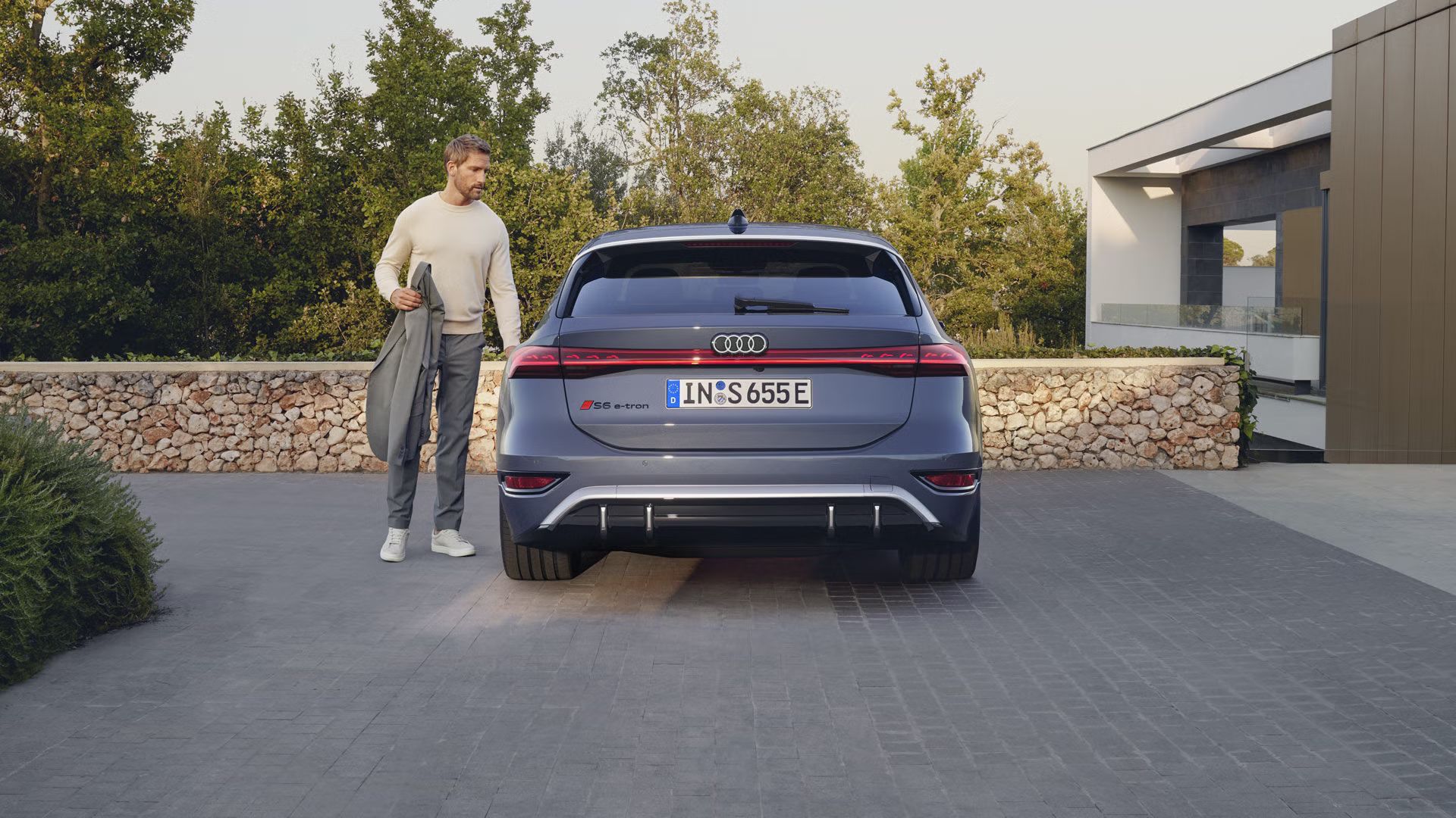 Heckprofil des S6 Avant e-tron, welcher auf Pflastersteinen vor einer Steinmauer und dem Garten eines modernen Hauses steht. Ein Mann mittleren Alters ist in Begriff, eine Tür zu öffnen.