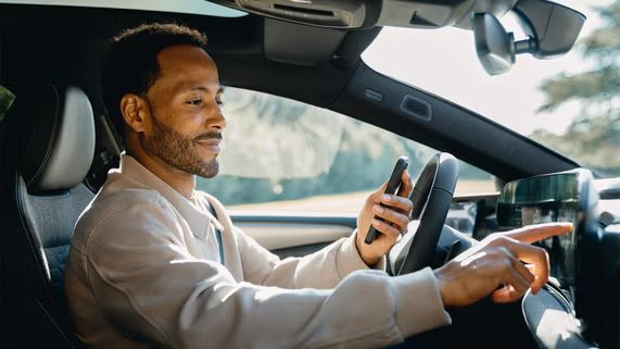 Ein Mann sitzt in einem Auto. In der linken Hand hält er ein Smartphone, mit der rechten Hand bedient er das digitale Mitteldisplay. 