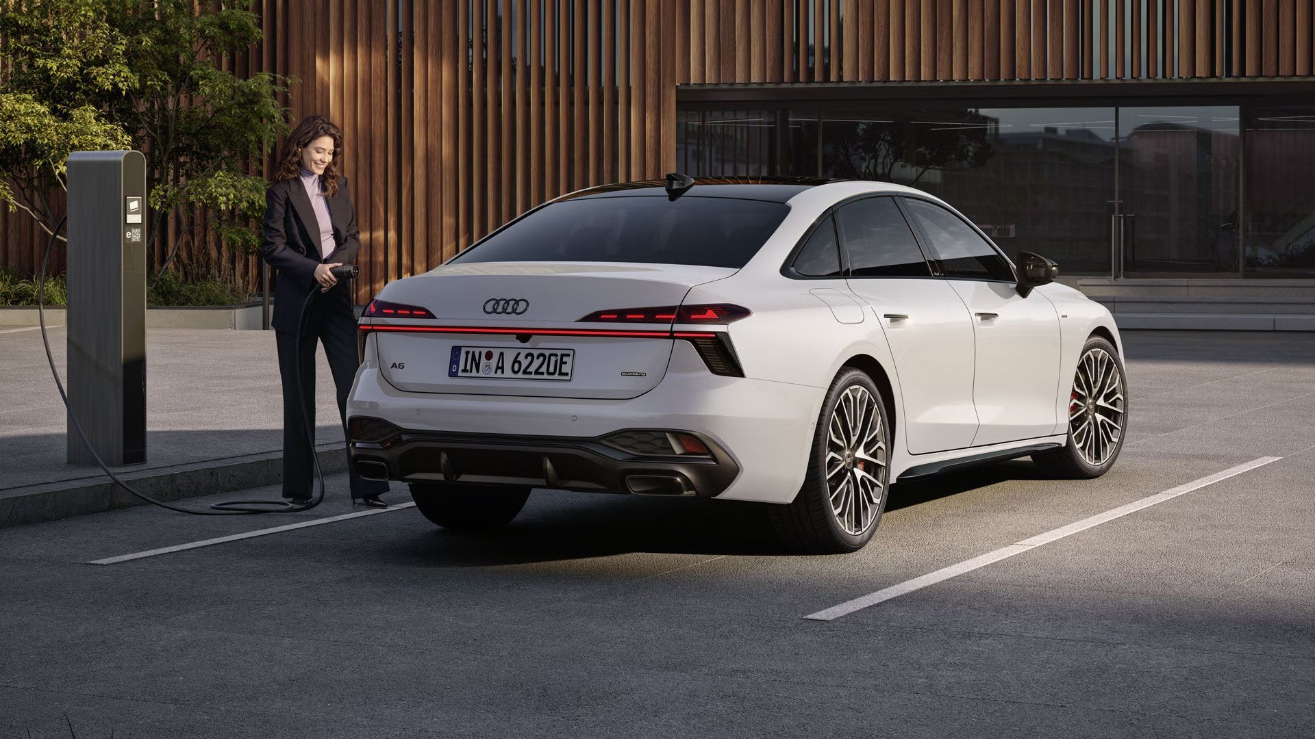 Die A6 Limousine e-hybrid in weiß steht auf einem Parkplatz vor einer Gebäudefassade. Eine junge Frau schließt die A6 Limousine e-hybrid an eine Ladestation an.