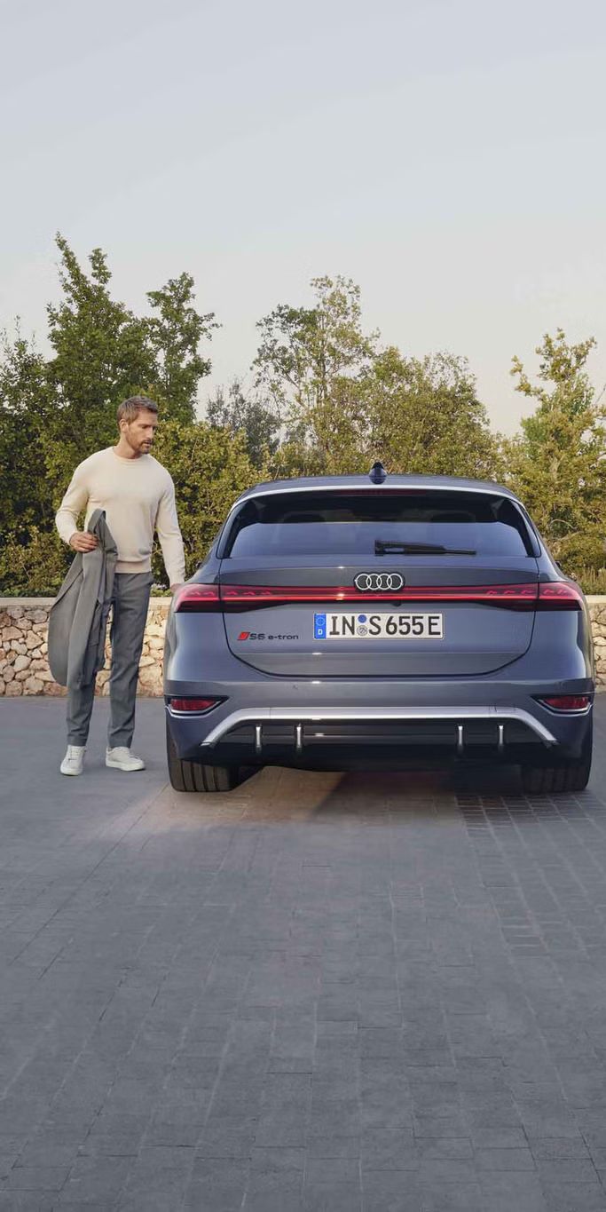 Heckprofil des S6 Avant e-tron, welcher auf Pflastersteinen vor einer Steinmauer und dem Garten eines modernen Hauses steht. Ein Mann mittleren Alters ist in Begriff, eine Tür zu öffnen.